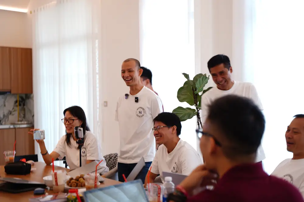 Học viên cười quanh bàn học, một người đang quay lại buổi học bằng điện thoại
